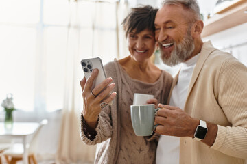 A happy couple laughs together while holding mugs and looking at a smartphone in a cozy kitchen.