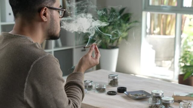 Asian man smoking a cannabis joint in a living room, with cannabis equipment and accessories on the table.