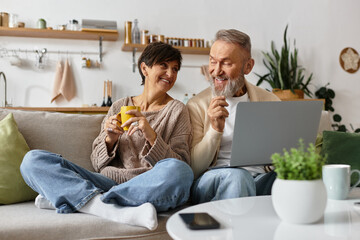 A joyful couple shares laughter and drinks while exploring a laptop in their warm home.
