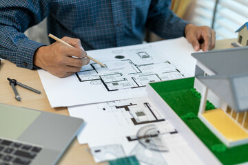 A man is holding a model of a house. The house is a small, two-story home with a slanted roof. The man is wearing a safety vest, which suggests that he is working on a construction site