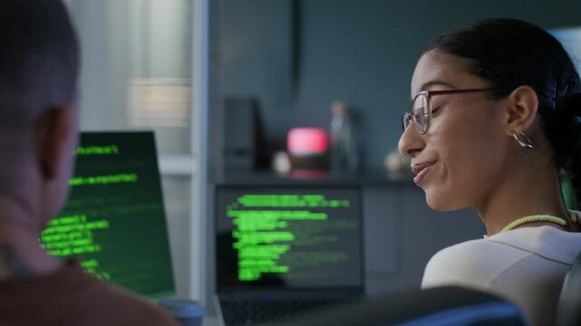 Back view shot of young female biracial programmer showing typing progress to her male Caucasian colleague and giving high five in modern office at night