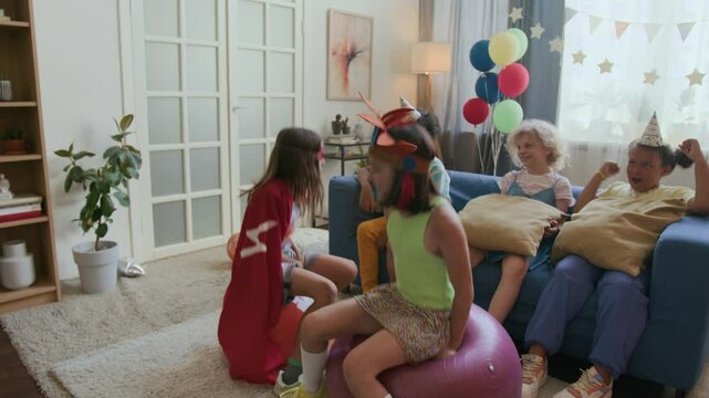 Two children jumping on hopper balls, three kids encouraging them relaxing on sofa at birthday party