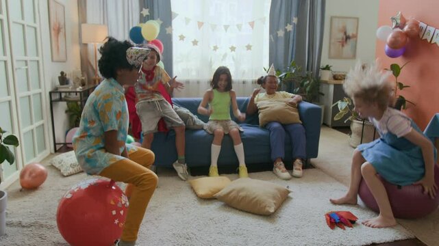 Two multi-ethnic kids bouncing on hopper balls while three friends cheering encouragingly in background during birthday party