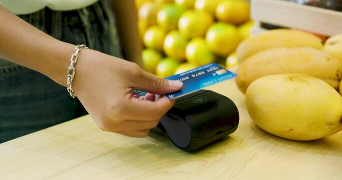 Asian woman making a cashless payment using a credit card and card reader in a fruit shop. Fresh yellow mangoes and fruits on the counter emphasize convenience and modern shopping. Credit card machine