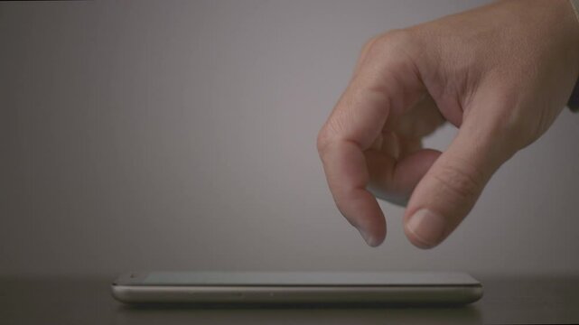 A close-up of a hand with fingers lightly pressing on a smartphone screen, displayed on a dark flat surface with a neutral background and subtle lighting emphasizing the interaction.