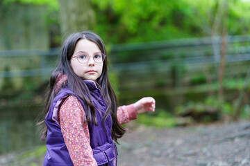 A small beautiful smiling long-haired dark-haired girl in glasses and a vest stands alone in the...