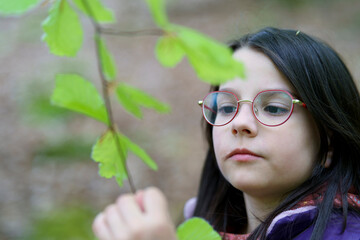 A small beautiful smiling long-haired dark-haired girl in glasses and a vest stands alone in the forest