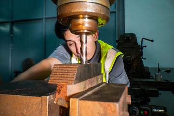 An industrial worker operating a drill press in a workshop. The focus is on the precision and...