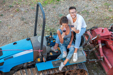 young couple of winemakers toasting with two glasses of wine sitting on an old crawler tractor at sunset