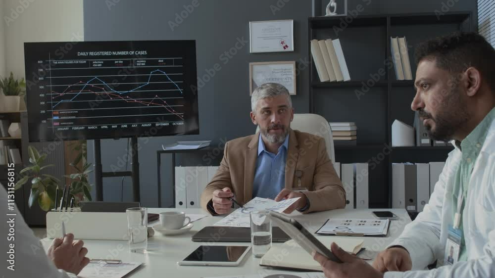 Poster middle-aged clinical manager showing documents and holding discussion with team of doctors sitting a