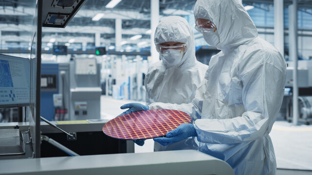 Semiconductor Production Process At Electronics Factory: Two Scientists In Sterile Suits Taking Out Silicon Wafer From Soldering Jet Printer And Checking It. Engineers Creating Hardware For Computers.