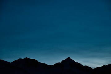 The mountain tops form a striking silhouette against a deep blue sky, their rugged outlines sharply defined. This tranquil scene evokes a sense of majesty and calm in nature's twilight.