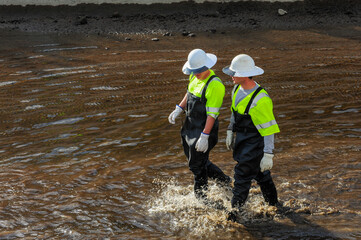 Water canal workers wearing rubber boots, black waders, and white hard hats walk through shallow...