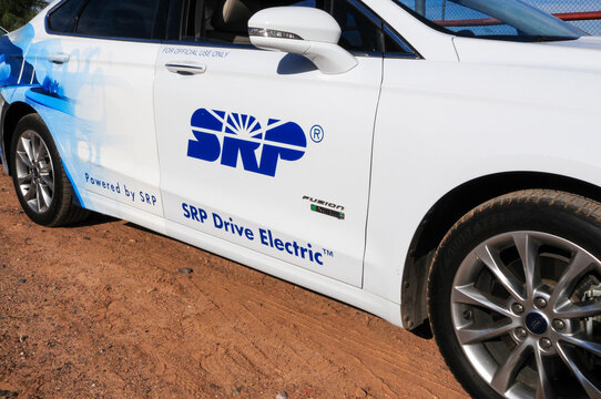 A partial view of a white Ford Fusion Energi plug-in hybrid, a four-door vehicle featuring the SRP (Salt River Project) logo and the trade name "SRP Drive Electric," parked along a dirt road