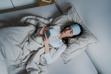 A woman laying on her back, checking messages on her phone with a cozy blanket wrapped around her.