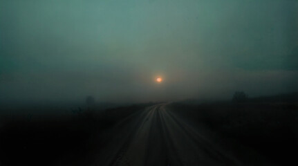 Fototapeta premium Mysterious Foggy Dirt Road at Dawn with Dim Sun Breaking Through Dense Green Mist, Creating Eerie Rural Atmosphere
