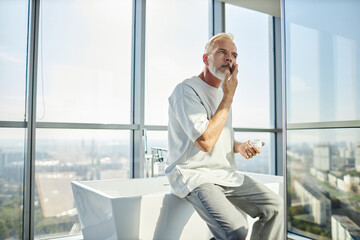 Medium shot of mature man in comfy home outfit applying nourishing cream on face taking care of skin while sitting on bathtub against big windows at home, copy space