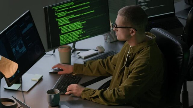 High angle shot of young Caucasian male programmer in eyeglasses wearing casualwear typing on pc computer sitting at computer desk with program codes on screen and sipping coffee in evening office