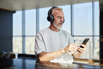 Medium shot of serious mature man with neat beard using smartphone listening to music in headphones while sitting at table in contemporary apartment, copy space