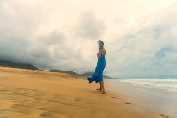 A woman in a flowing blue dress running barefoot on the sandy Playa de Cofete beach in Fuerteventura, Canary Islands. The dramatic cloudy sky, golden sand, and ocean waves 