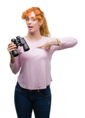 Young redhead woman looking through binoculars with surprise face pointing finger to himself