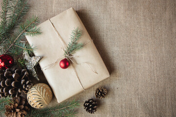 A gift wrapped in paper is placed on a burlap background, surrounded by Christmas decorations. Copy space. Vintage style.