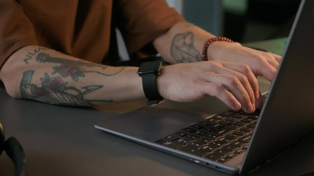 Slowmo close up shot of male programmer with tattooed arms wearing casualwear working on laptop in office at night
