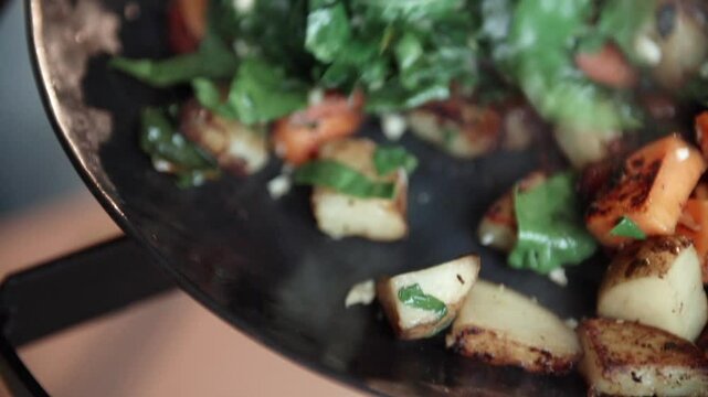Closeup shot of a wooden spatula flipping and mixing potatoes and swiss chard in a hot steel pan