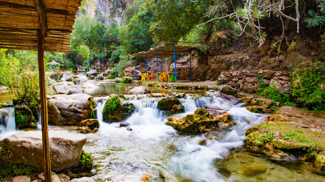 A clear river flows through lush green rocks in Akchour, Chefchaouen. Colorful chairs under a rustic shelter create a peaceful and picturesque setting by the water.
