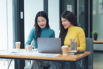 Obraz premium Collaborative Work: Two young women, dressed in casual attire, work together on a project, demonstrating focus and teamwork. They are engaged in a discussion, looking at a laptop, and taking notes.