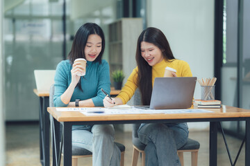 Collaborative Coffee Break: Two smiling Asian female colleagues enjoy a coffee break while working...