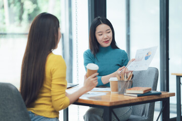 Fototapeta premium Businesswomen Discussing Data: Two young Asian businesswomen engage in a productive discussion over coffee, reviewing financial charts and graphs. A scene of collaboration and professional growth. 