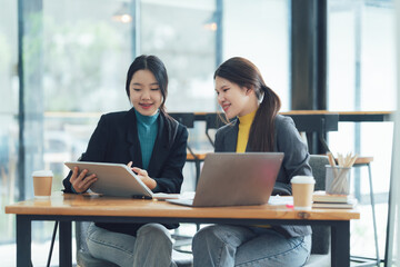 Collaborative Businesswomen: Two young Asian businesswomen engage in a productive discussion using a tablet and laptop, fostering teamwork and innovation in a modern office setting.