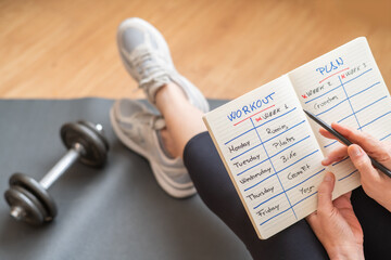 Mature Fitness Woman Sitting On Yoga Mat With Dumbbells, Writing Morning Exercise Schedule On...