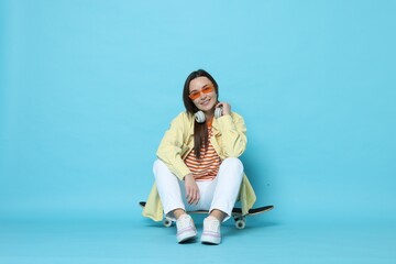 Fototapeta premium Smiling woman sitting on skateboard against light blue background