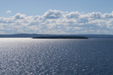Naklejka premium Vom Sonnendeck der Fähre von Oslo nach Kopenhagen ist eine Insel im Oslofjord zu sehen. Sie liegt an diesem Sommertag im Gegenlicht, sodass die Wellen schimmern. Im Hintergrund sind Wolken sichtbar.