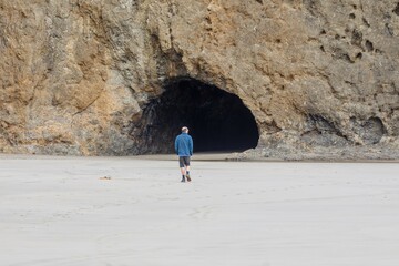 A lone hiker walks along a sandy beach towards a large cave in the cliff face. Exploring the...