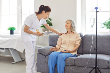 Smiling friendly young nurse or caregiver checking condition of senior elderly woman patient sitting on sofa and receiving intravenous IV drip infusion and vitamin injection therapy at nursing home.