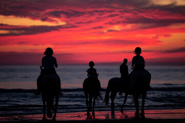Obraz premium Contraluz de personas montando a caballo en la playa en el atardecer