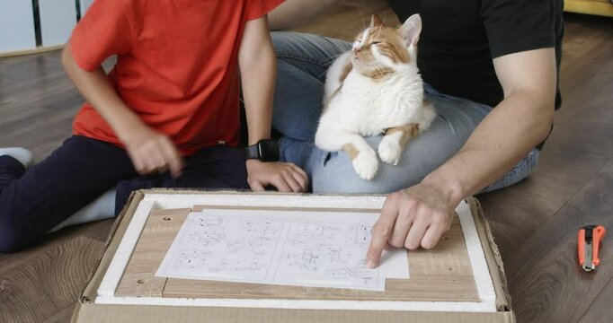 Dad and son sit looking at instructions for assembling nightstand. Cat lies on father crossed legs. Man and young boy assemble furniture