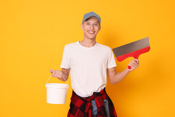 Portrait of young decorator with putty knife and bucket on orange background