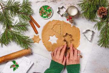 Process of cooking Christmas gingerbread cookies. Rolled raw dough with cookie cutters, cinnamon and pine branches on light stone table. Seasonal pastry. Woman making cookies. Top view.