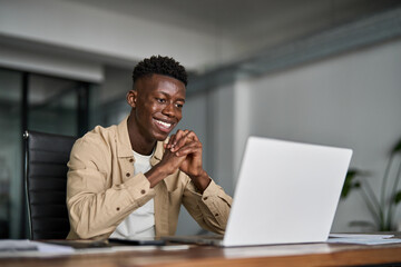 Happy young African ethnic business man looking at laptop working in office having virtual meeting. Smiling professional businessman trader using computer feeling satisfied with market growth at work.