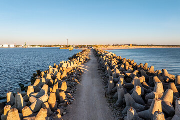 Obraz premium A long pathway bordered by concrete tetrapods extends into calm water, with a small lighthouse and industrial buildings visible under a clear sky.