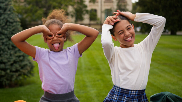 Happy little girls two kids schoolgirls children school education students pupils classmates multiracial African Indian female friends laugh have fun eyes circles grimace play game kidding outdoors