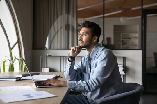 Serious Indian businessman deep in thought, reflecting on work-related issue or strategic decision seated at desk with laptop, looks into distance, analyzing data, considering new business opportunity