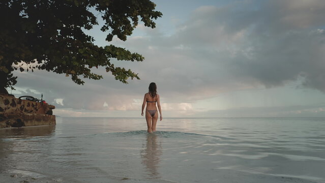 Young woman in a bikini is enjoying a relaxing evening on a tropical beach, walking into the calm ocean water as the sun sets over the horizon