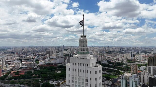 Vista a&eacute;rea do centro urbano de S&atilde;o Paulo, destacando o Farol Santander com a bandeira da cidade ao topo, cercado por pr&eacute;dios hist&oacute;ricos e modernos em harmonia