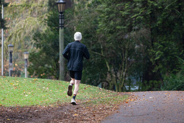 Fototapeta premium A man in a gray sweatshirt and black shorts jogs along a damp park trail in Portland, OR. Surrounded by greenery and fresh air, the peaceful setting makes for a healthy outdoor activity