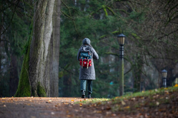 A person wearing a long coat, gray hat, and a red hiking backpack walks through a serene park trail...